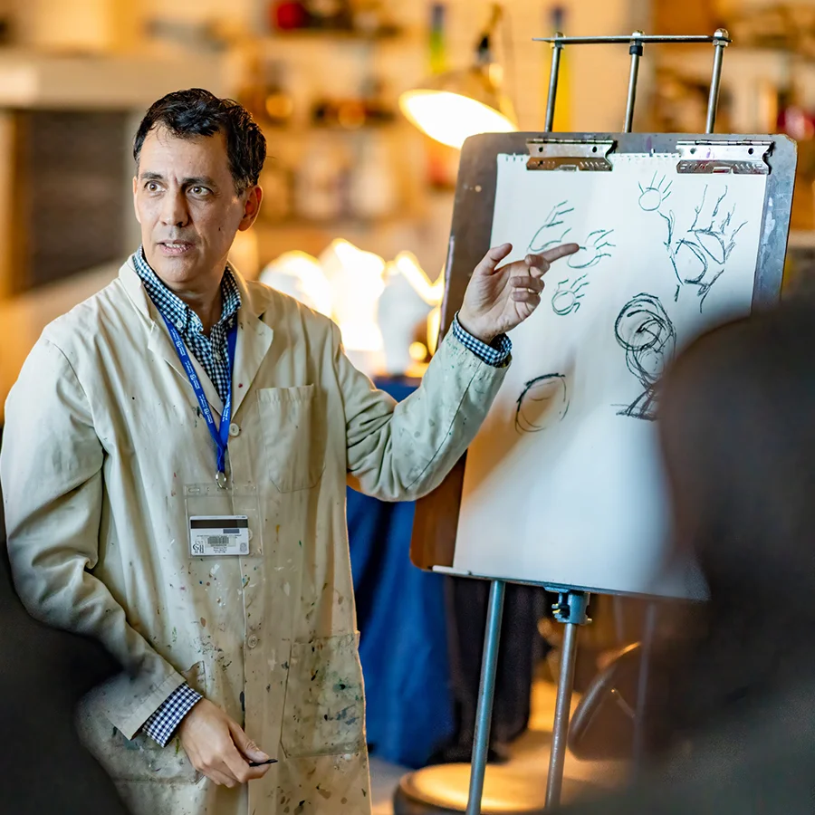An art professor teaches while gesturing towards an easel with a sketch.